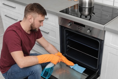 Photo of Young man cleaning oven with rag and detergent in kitchen