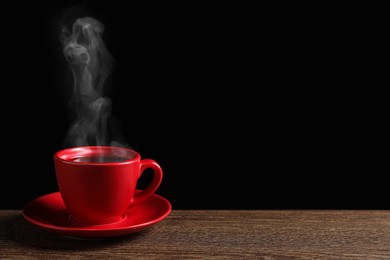 Red cup with hot steaming coffee on wooden table against black background, space for text