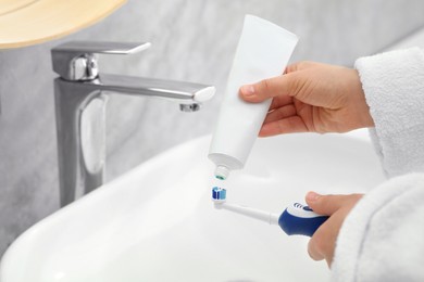 Photo of Woman squeezing toothpaste from tube onto electric toothbrush above sink in bathroom, closeup