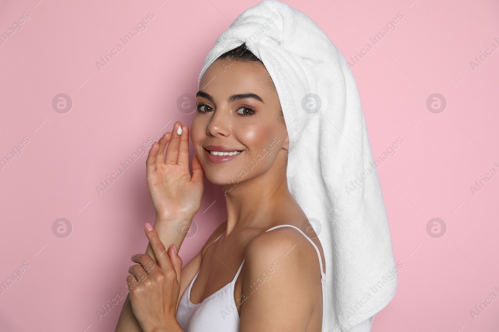 Photo of Beautiful young woman with towel applying cream on face against pink background