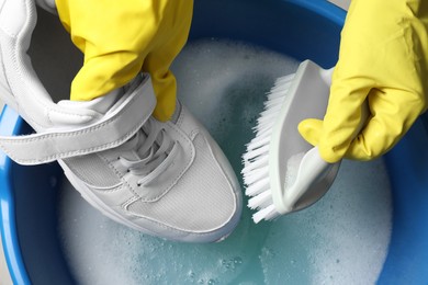 Woman with gloves and brush cleaning stylish sneakers in wash basin, top view