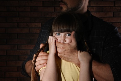 Adult man covering scared little girl's mouth near brick wall, closeup. Child in danger