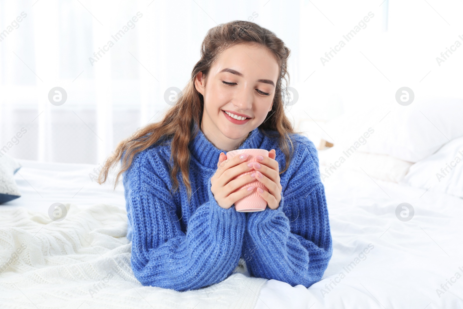Photo of Beautiful teenage girl in warm cozy sweater with cup of hot drink on bed at home