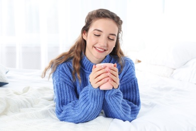 Photo of Beautiful teenage girl in warm cozy sweater with cup of hot drink on bed at home
