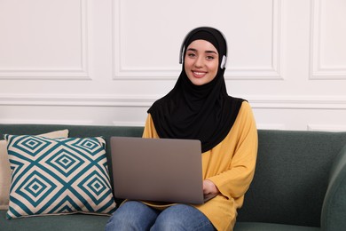 Photo of Muslim woman in hijab and headphones using laptop on sofa indoors