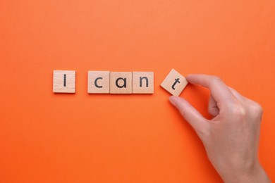 Photo of Motivation concept. Woman changing phrase from I Can't into I Can by removing wooden square with letter T on orange background, top view