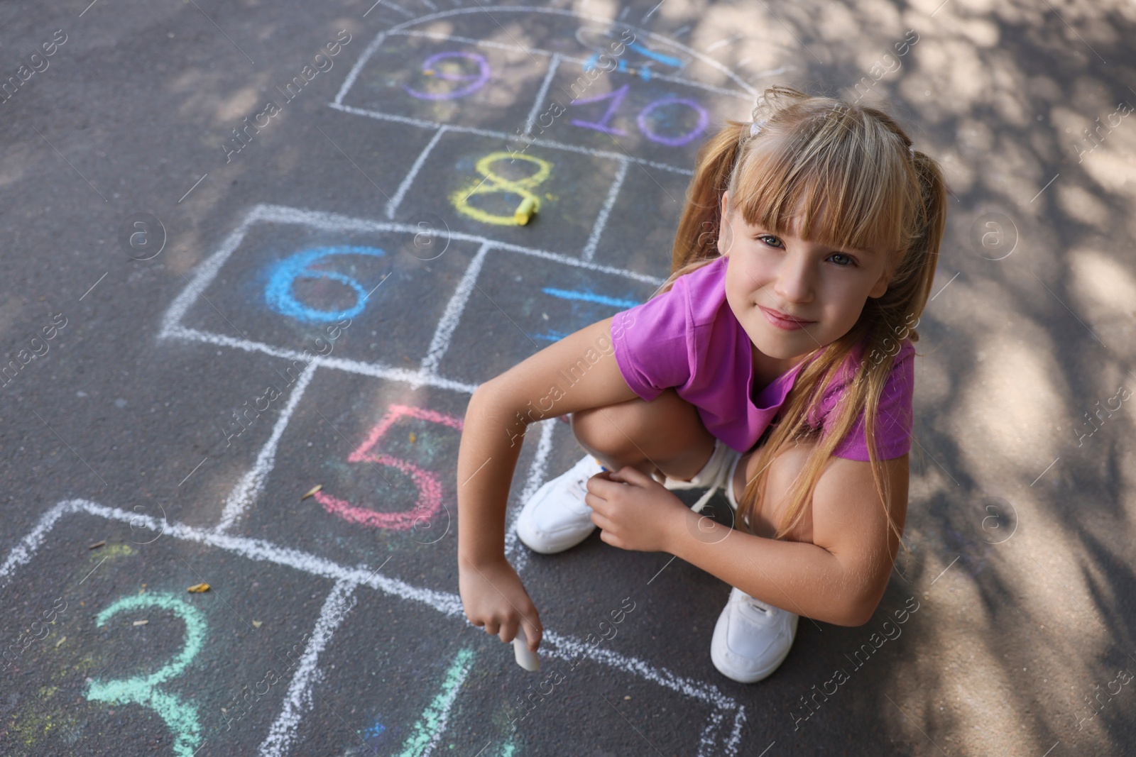 Photo of Little girl drawing hopscotch with chalk on asphalt outdoors. Happy childhood