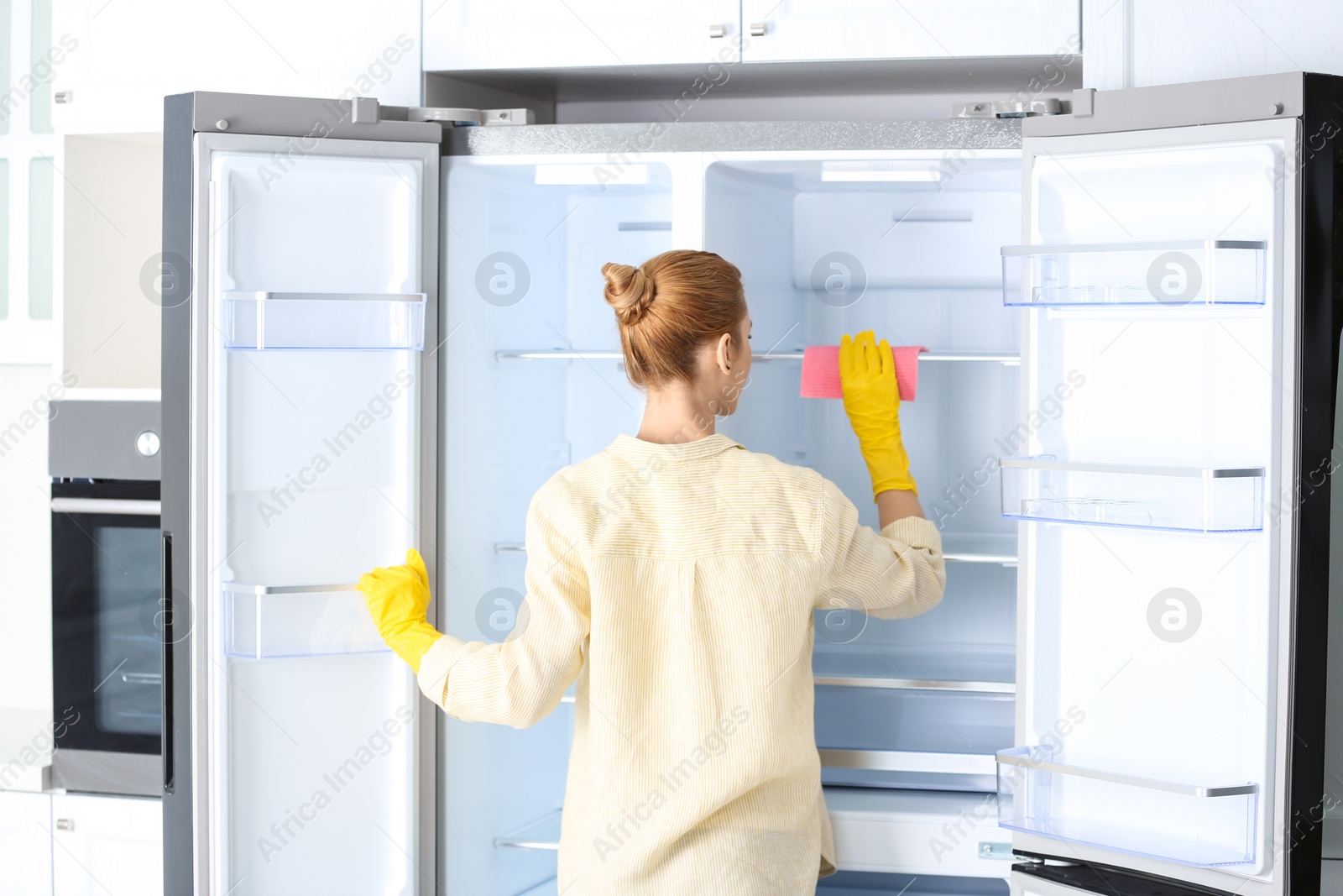 Photo of Woman in rubber gloves cleaning empty refrigerator at home, back view