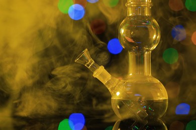 Photo of Closeup view of glass bong with smoke against blurred lights, toned in yellow. Smoking device