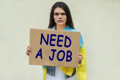 Unemployed due to war. Sad woman with flag of Ukraine holding cardboard sheet with phrase Need A Job on light background