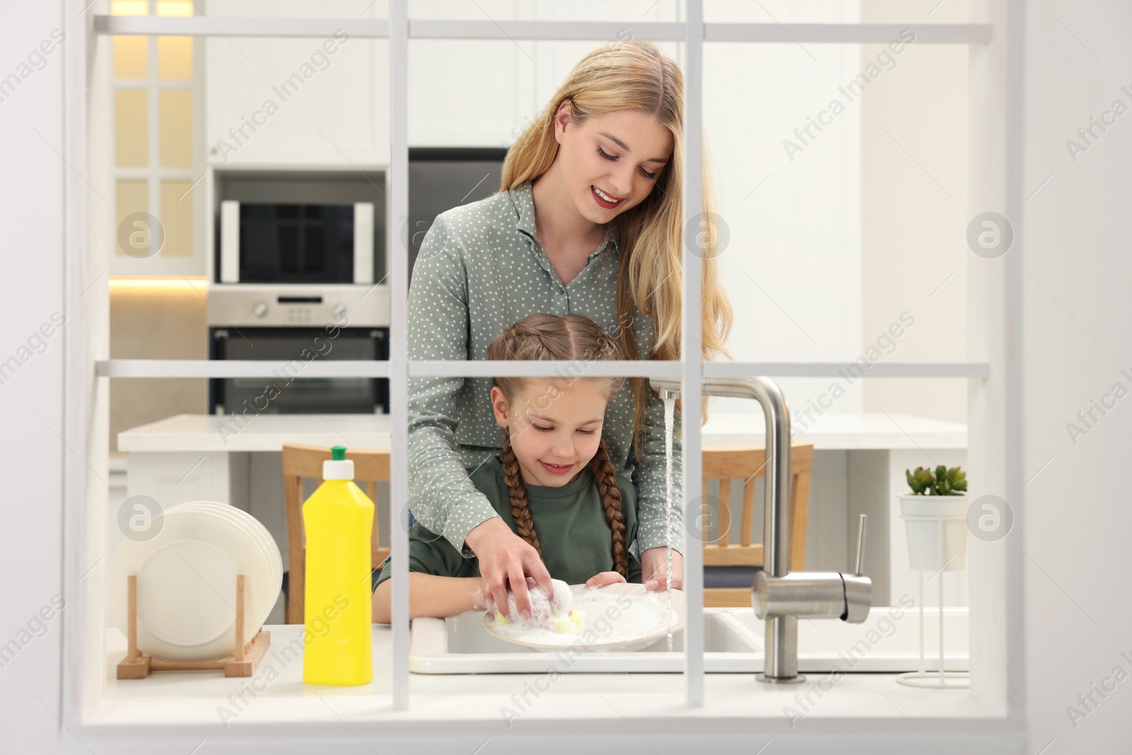 Photo of Mother and daughter washing plate above sink in kitchen, view from outside