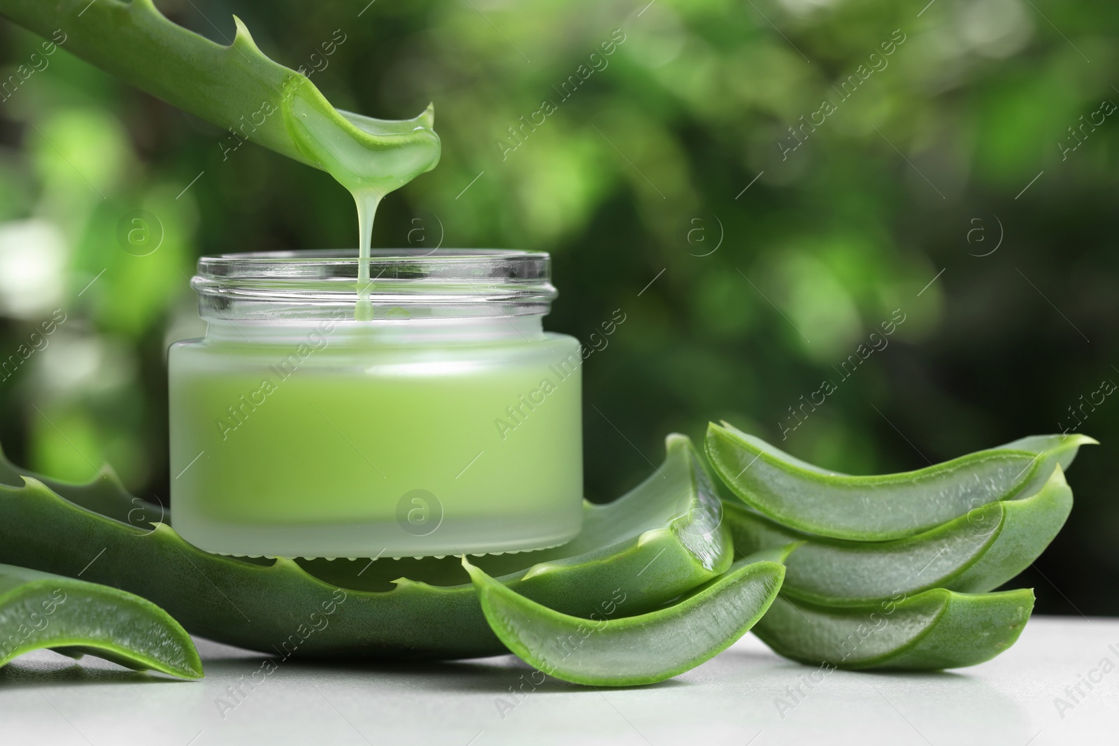 Photo of Aloe vera juice dripping from leaf into jar with cream on white table against green blurred background, closeup