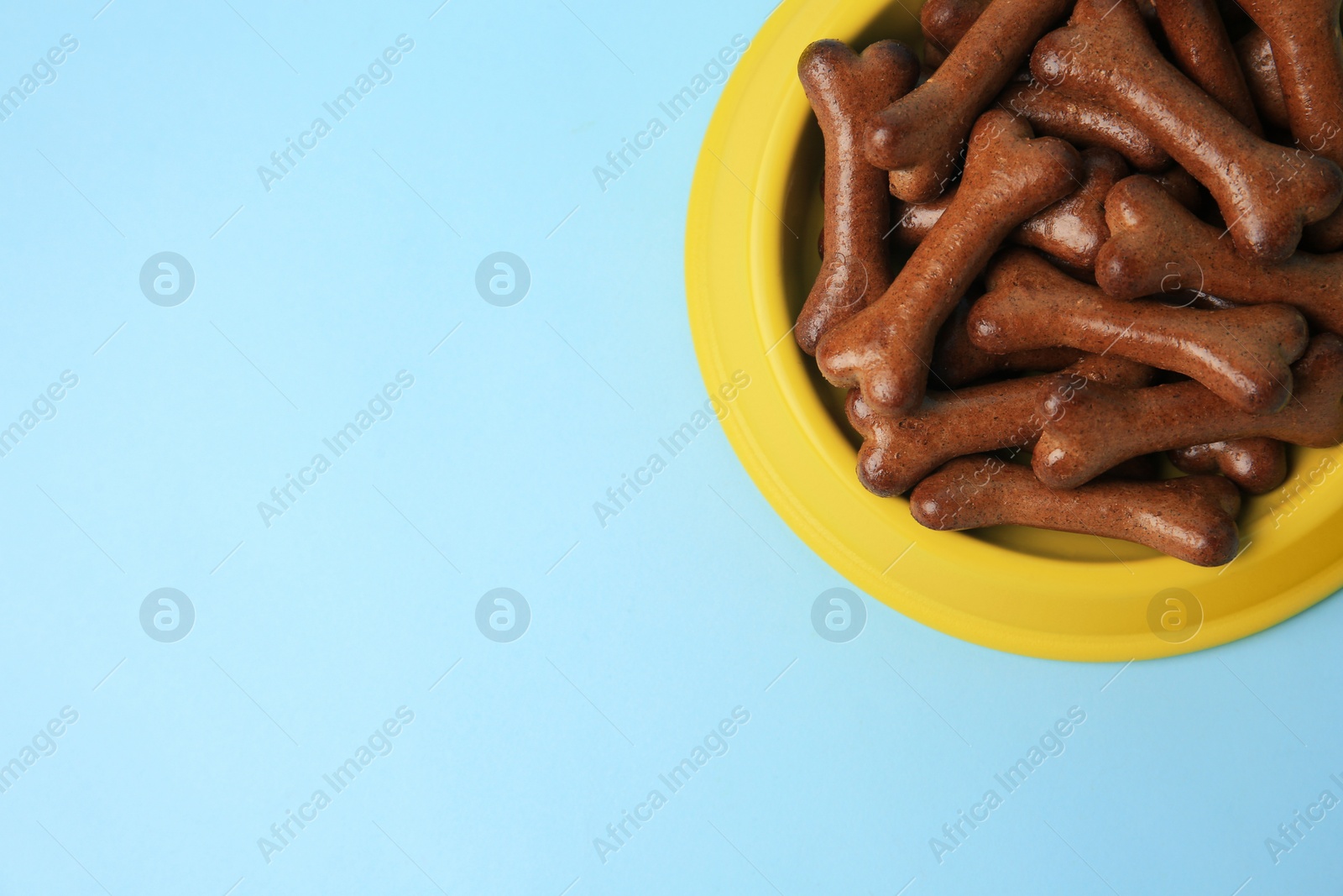 Photo of Yellow bowl with bone shaped dog cookies on light blue background, top view. Space for text