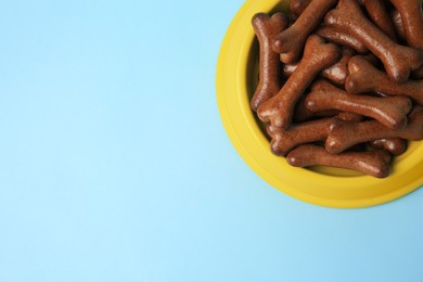 Photo of Yellow bowl with bone shaped dog cookies on light blue background, top view. Space for text