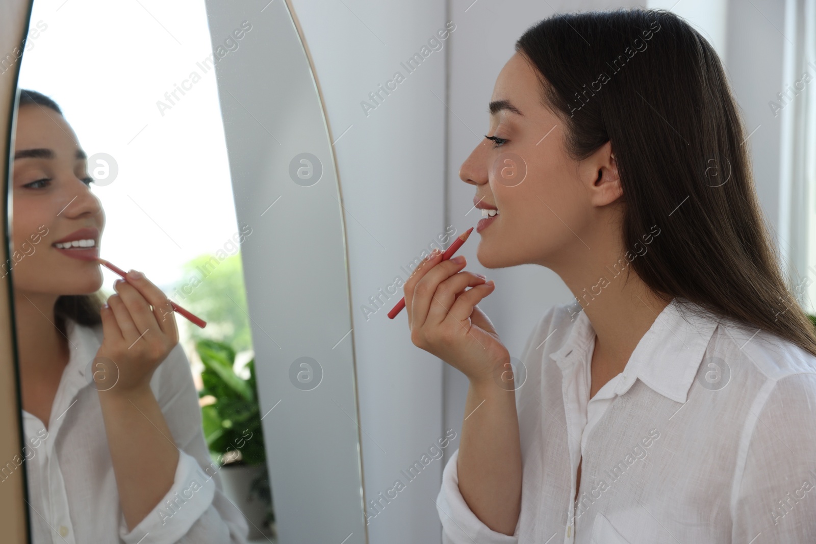 Photo of Beautiful young woman applying cosmetic pencil on lips near mirror indoors