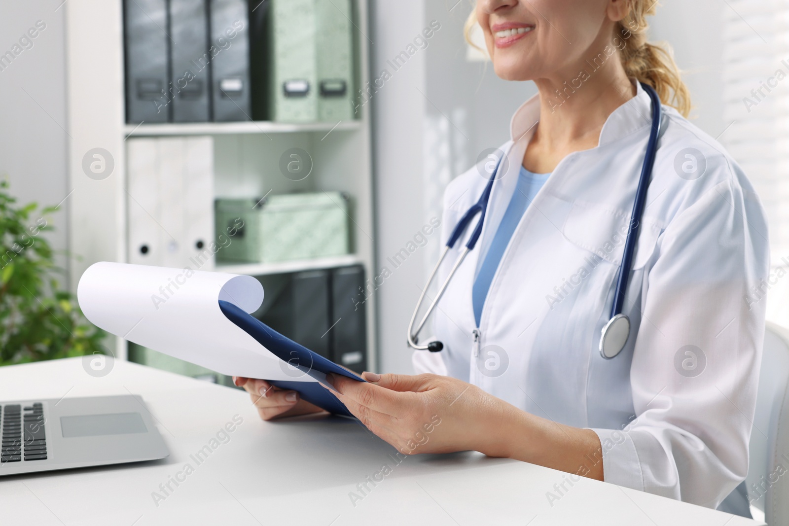 Photo of Doctor with laptop and clipboard at white table in clinic, closeup. Patient consultation