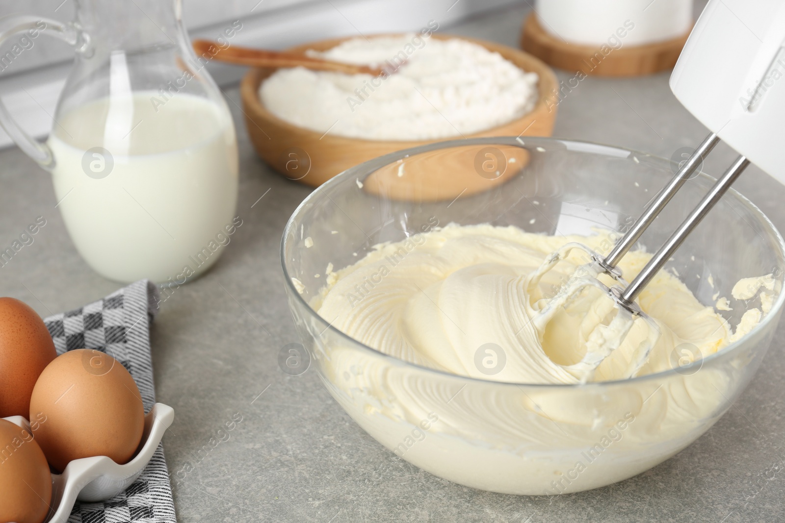 Photo of Whipping white cream in glass bowl with mixer on light grey table, closeup