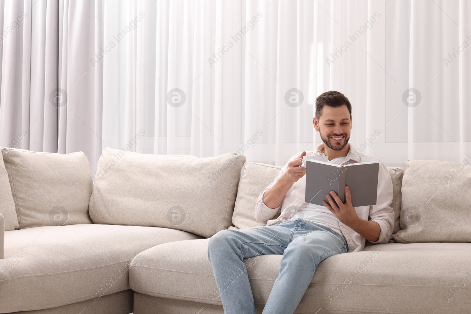 Photo of Happy man drinking coffee while reading book on sofa near window with beautiful curtains at home in living room