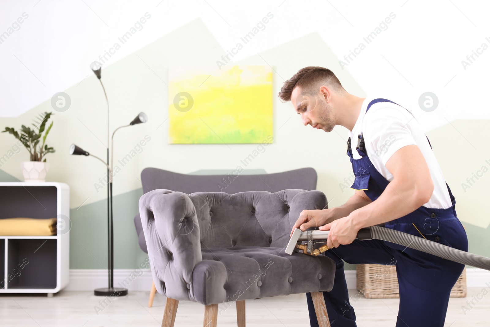 Photo of Male worker removing dirt from armchair with professional vacuum cleaner indoors