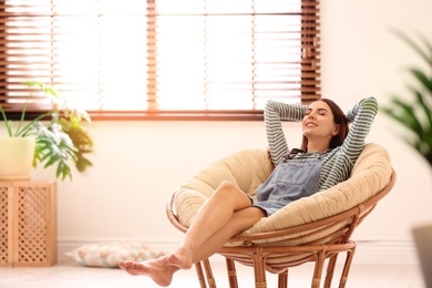 Photo of Young woman relaxing in papasan chair near window with blinds at home. Space for text