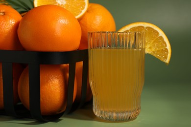 Fresh oranges in metal basket and glass of juice on green background, closeup