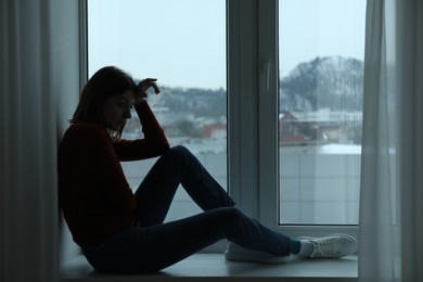 Photo of Sad young woman sitting on windowsill near window at home
