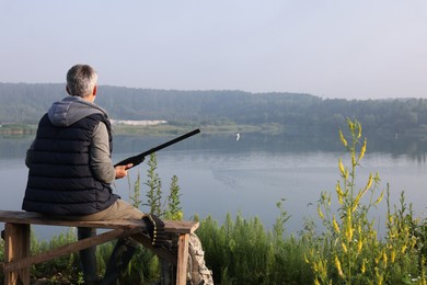 Man with hunting rifle sitting on wooden bench near lake outdoors, back view. Space for text