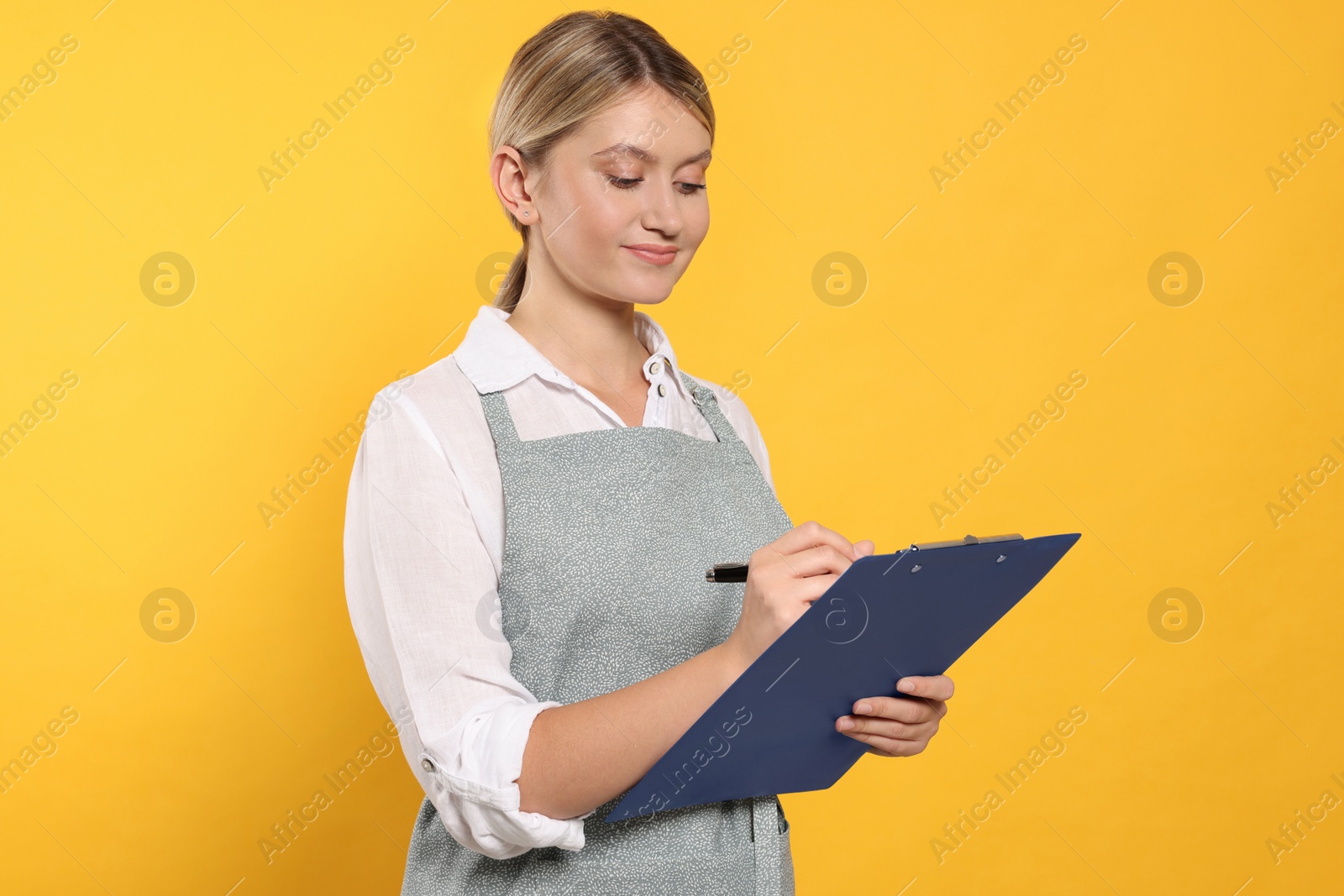 Photo of Beautiful young woman in clean apron with clipboard on orange background