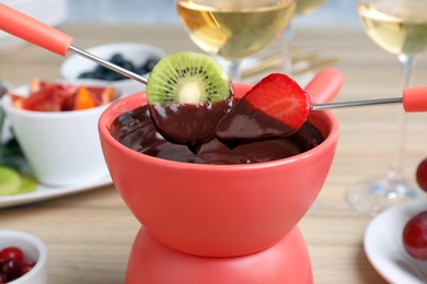 Photo of Dipping slices of strawberry and kiwi into pot with chocolate fondue at table, closeup