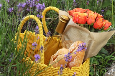 Yellow wicker bag with beautiful roses, bottle of wine and baguettes in lavender field, closeup