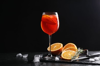 Glass of tasty Aperol spritz cocktail with orange slices, ice cubes and rosemary on table against black background