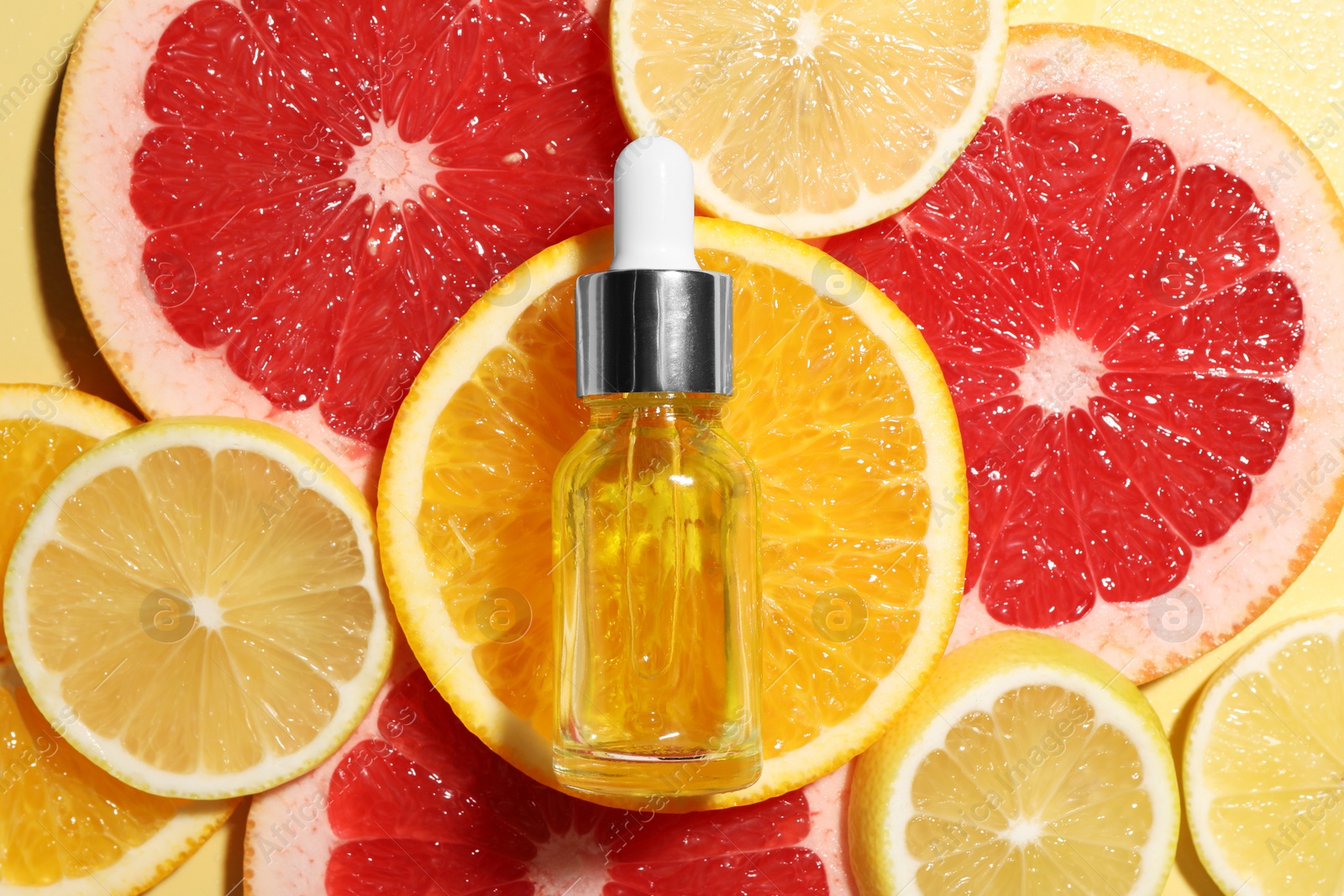 Photo of Bottle of cosmetic serum and sliced citrus fruits on yellow background, flat lay