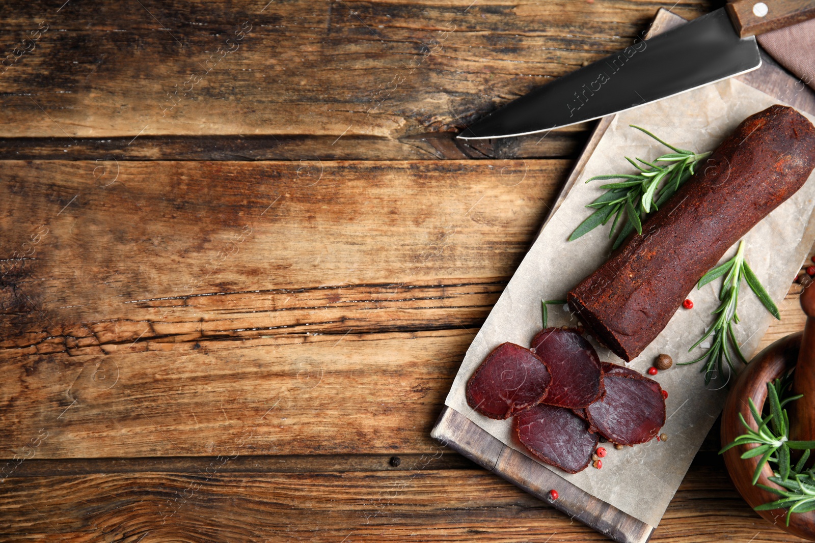 Photo of Delicious dry-cured beef basturma with rosemary and peppercorns on wooden table, flat lay. Space for text