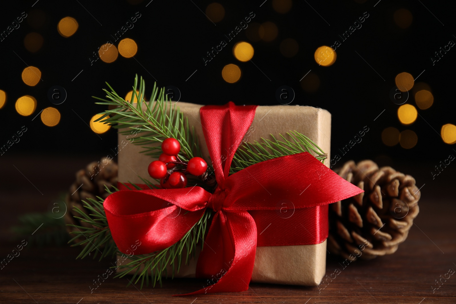 Photo of Gift box with fir tree branches and pine cones on wooden table against blurred festive lights