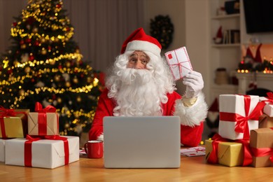 Photo of Santa Claus with letter surrounded by Christmas gifts at table in room