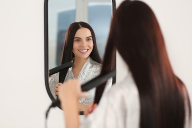 Photo of Beautiful happy woman using hair iron near mirror indoors