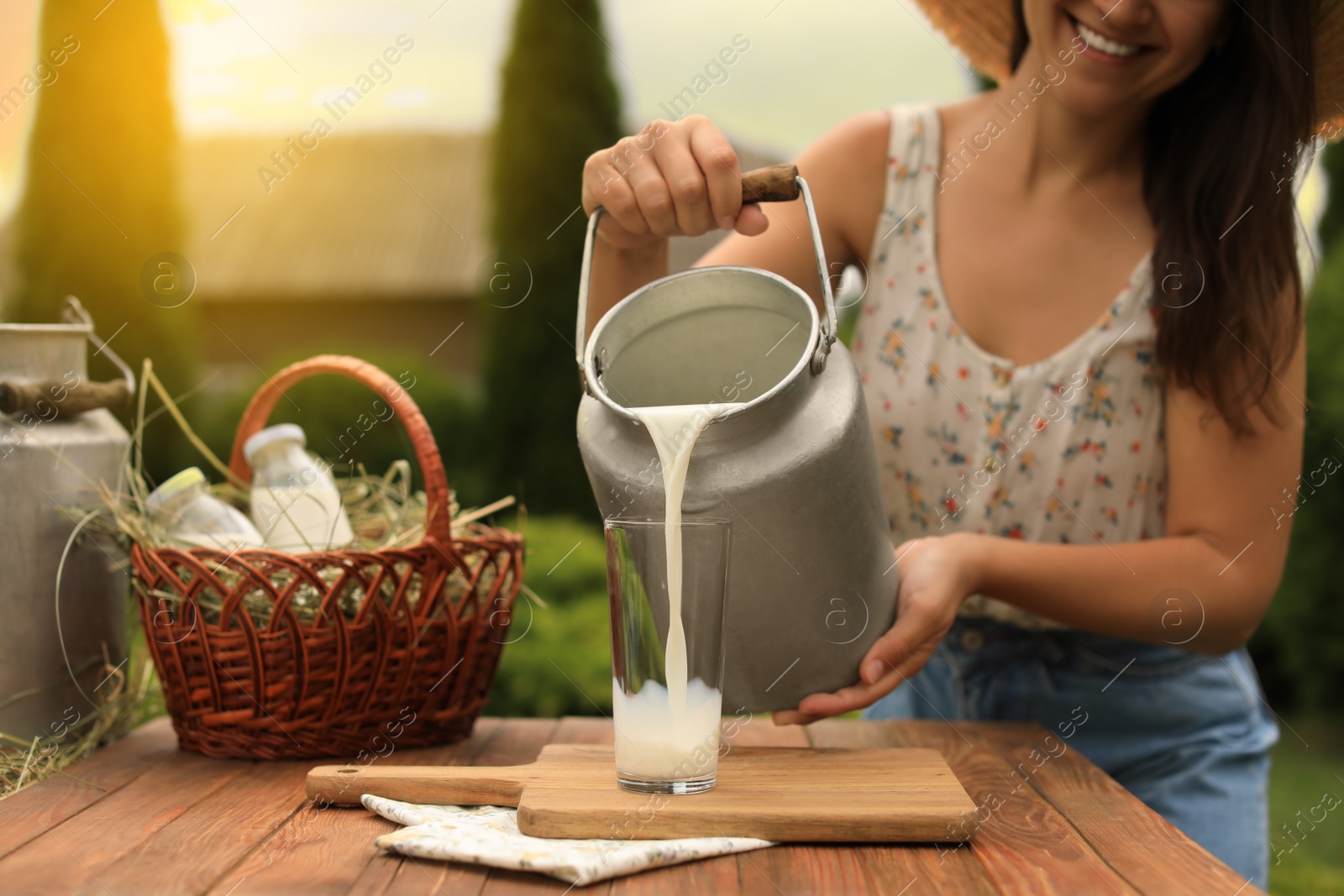 Photo of Smiling woman pouring fresh milk from can into glass at wooden table outdoors, closeup