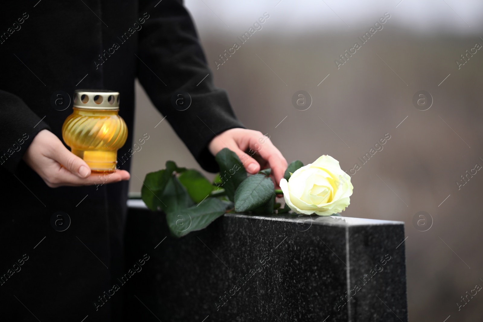 Photo of Woman with candle and rose near black granite tombstone outdoors, closeup. Funeral ceremony