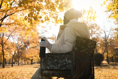 Woman with thermos sitting in camping chair outdoors on autumn sunny day