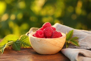 Tasty ripe raspberries in bowl and green leaves on wooden table outdoors