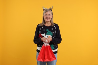 Photo of Happy senior woman in Christmas sweater and deer headband with shopping bags on orange background