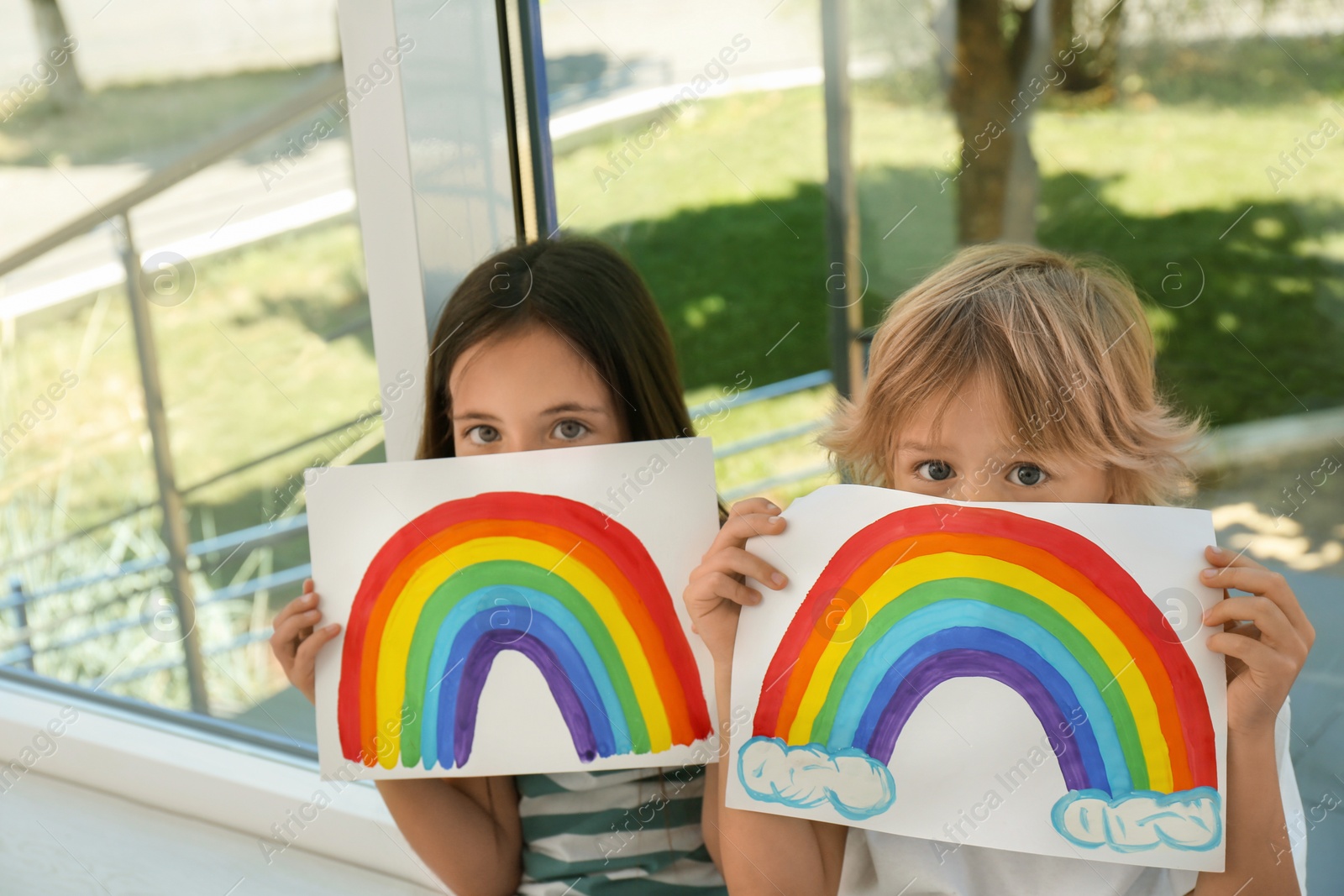 Photo of Little children holding rainbow paintings near window indoors. Stay at home concept