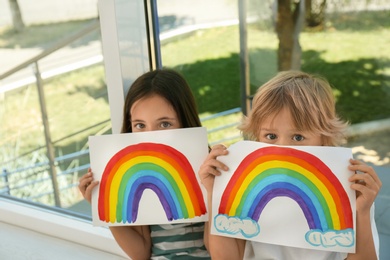 Photo of Little children holding rainbow paintings near window indoors. Stay at home concept