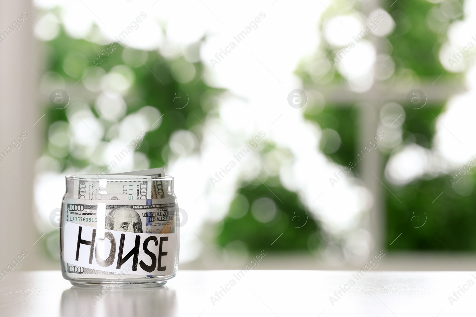 Photo of Glass jar with money and word HOUSE on table against blurred background, space for text