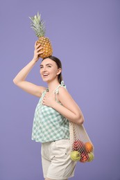 Photo of Woman with string bag of fresh fruits holding pineapple above her head on violet background