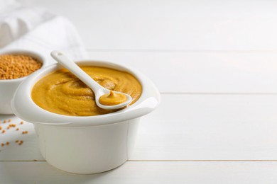 Photo of Spoon and bowl of tasty mustard sauce on white wooden table, space for text