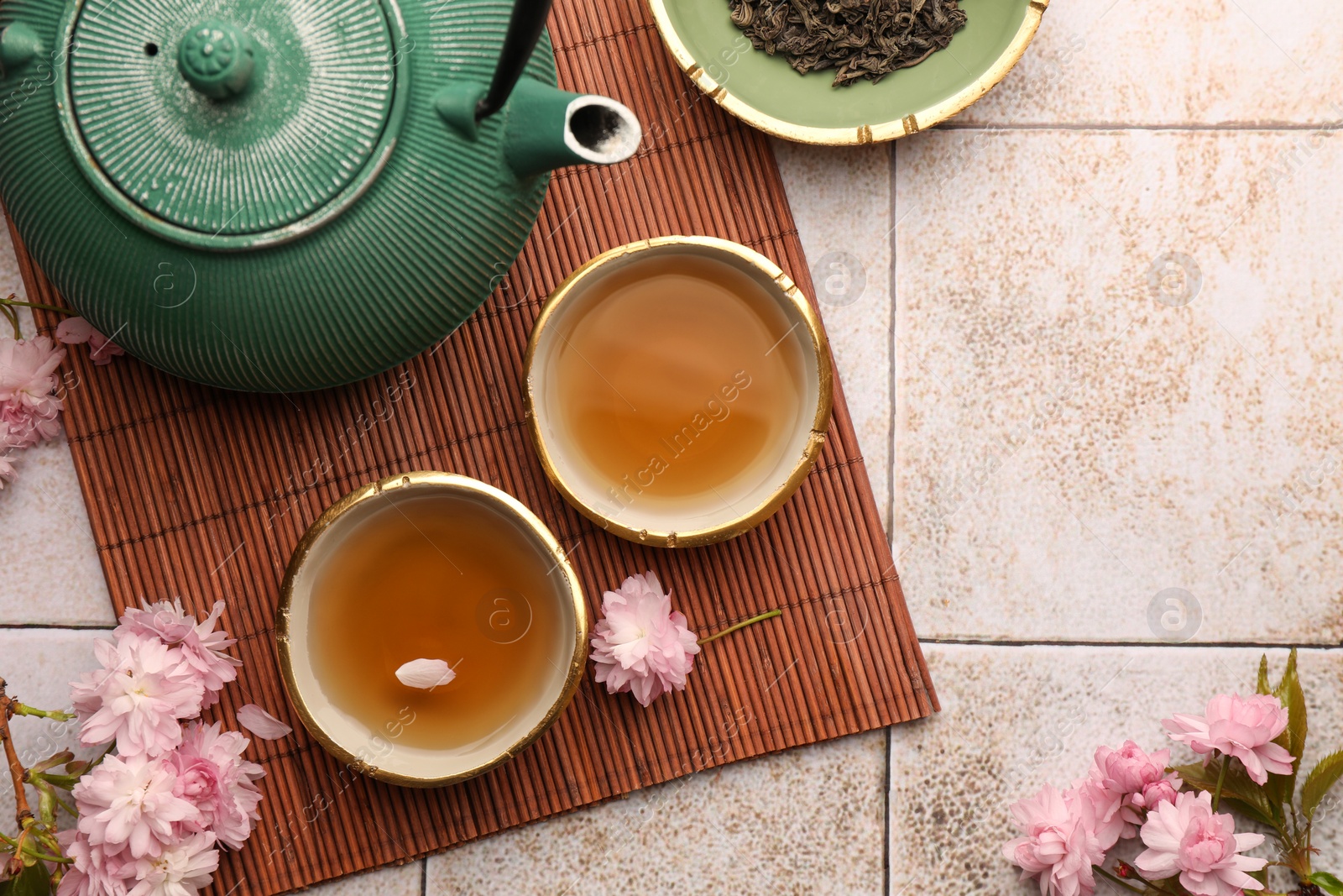 Photo of Traditional ceremony. Cup of brewed tea, teapot, dried leaves and sakura flowers on tiled table, flat lay with space for text