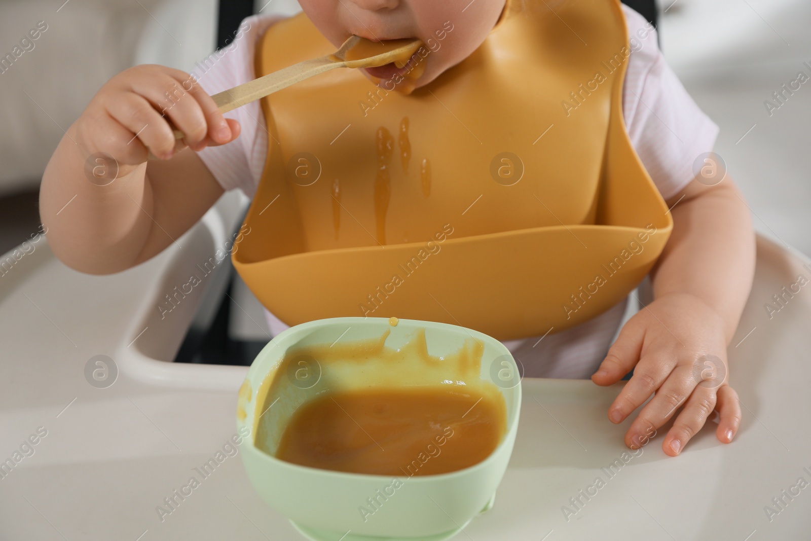 Photo of Cute little baby wearing bib while eating indoors, closeup