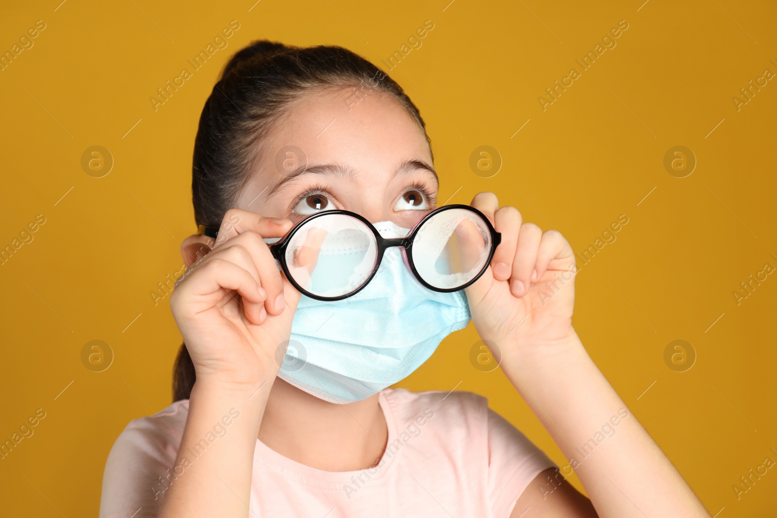 Photo of Little girl wiping foggy glasses caused by wearing medical face mask on yellow background. Protective measure during coronavirus pandemic