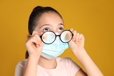 Little girl wiping foggy glasses caused by wearing medical face mask on yellow background. Protective measure during coronavirus pandemic